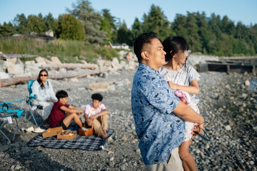 A family enjoying a summer evening on a rocky beach in Surrey BC, with a father holding his daughter while children and a woman relax on a blanket in the background